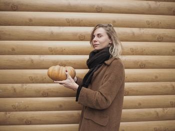 Side view of woman holding pumpkin while standing by wooden wall