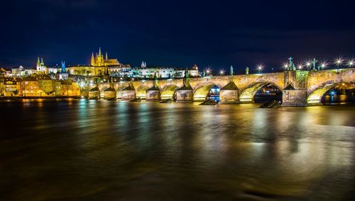 Illuminated old arch bridge over river in town against sky at night