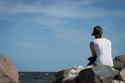 Rear view of woman sitting on rock while looking at sea against sky