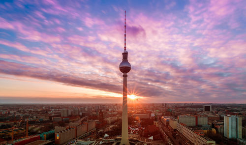 Communications tower in city against cloudy sky at sunset