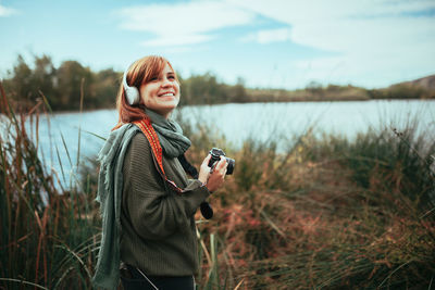 Young woman photographing while standing by lake against sky
