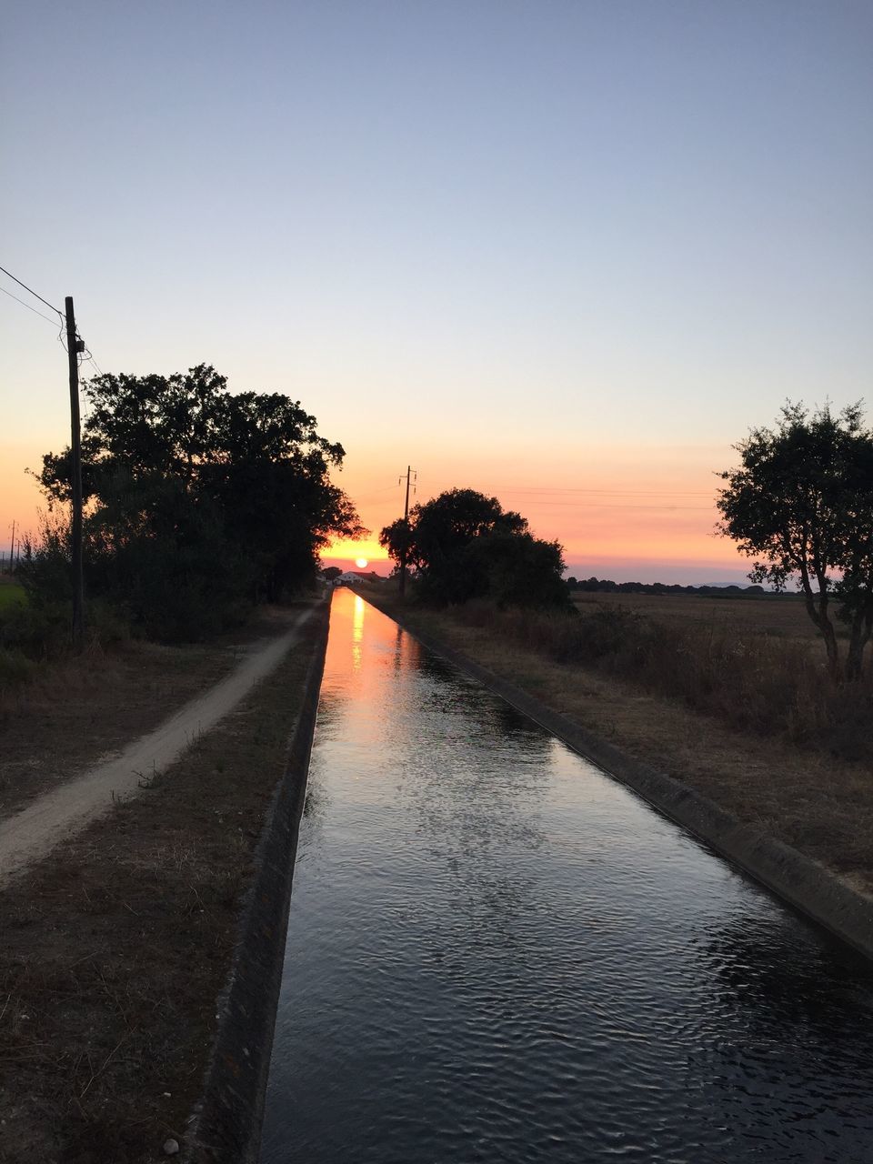 sunset, tree, the way forward, road, transportation, water, clear sky, vanishing point, street, copy space, diminishing perspective, orange color, tranquil scene, tranquility, footpath, outdoors, day, blue, sky, empty road, scenics, nature, country road, non-urban scene, sea, sun, long, solitude, beauty in nature