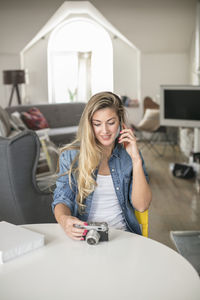 Young woman holding camera talking on mobile phone while sitting by table at home