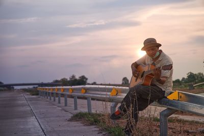 Man sitting on railing against sky during sunset and playing the guitar