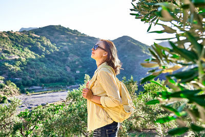 Woman stending in olive grove, enjoying mountain view, traveling with backpack 