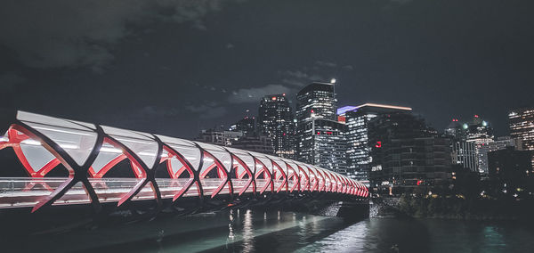 Illuminated bridge over river by buildings against sky at night