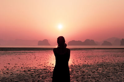 Rear view of silhouette woman standing on beach against sky during sunset