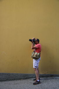 Side view of man photographing against yellow wall