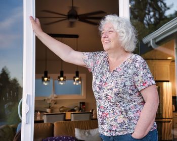 A happy elderly grey-haired woman smiles at the doorway of her house.