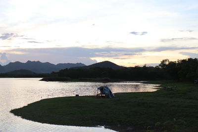 People sitting on land by lake against sky during sunset