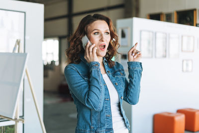 Charming emotional young woman designer artist in jeans jacket using mobile phone in art gallery