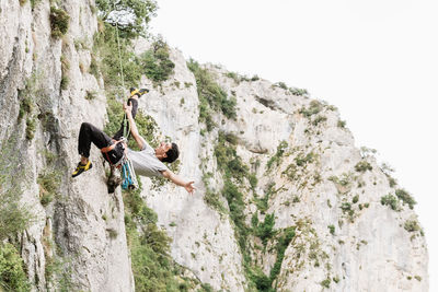 Full length of cheerful male hiker shouting while rappelling on rocky cliff against sky