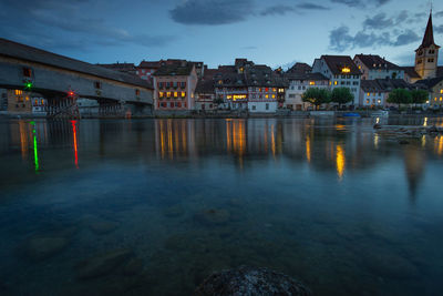 Illuminated buildings by river against sky at dusk