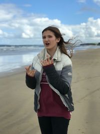 Portrait of young woman gesturing horn sign while standing at beach against sky