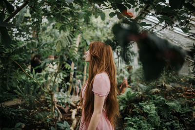Side view of of redhead female standing among tropical palm and plants in glasshouse