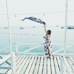 Woman holding scarf while standing on pier by sea