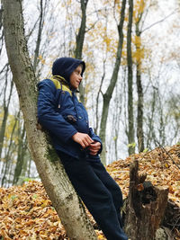 Boy stands leaning under a tree in the autumn forest and looks away