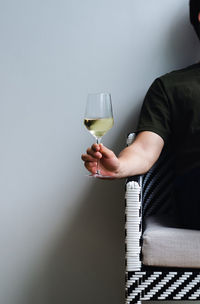 A glass of white wine holding by a person sitting on chair with shadow on white wall background.