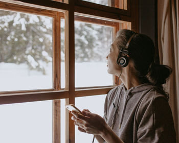 Woman in country house at the window with headphones and mobile phone