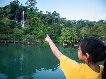 Rear view of woman pointing at waterfall by lake against trees and sky