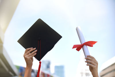 Woman holding diploma and mortarboard against sky