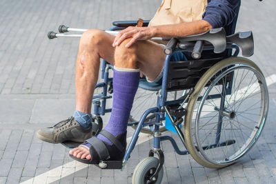 Middle-aged caucasian man sits in a wheelchair with a cast on his broken leg, holding crutches in