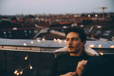 Thoughtful young man sitting on illuminated terrace in city during party