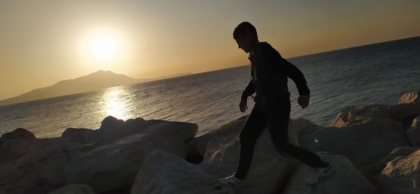 Man on rock at beach against sky during sunset