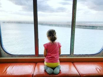 Rear view of girl looking at sea through window while sitting on seat at home