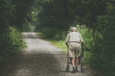 Rear view full length of senior man with mobility walker on road amidst trees