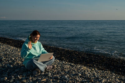 Young woman sitting at beach against sky