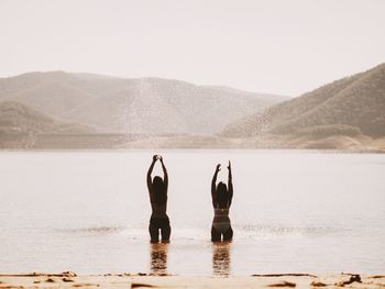 Rear view of friends with arms outstretched standing on beach by mountains against clear sky