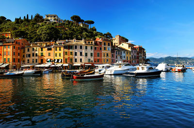 Boats moored in sea by buildings against blue sky