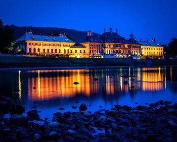 Illuminated buildings by lake against blue sky at dusk