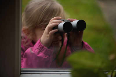 Close-up of girl looking through binoculars seen through window