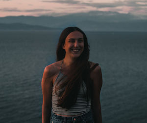 Smiling young woman standing at sea during sunset