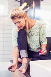 Full length of young woman painting toe nails while sitting on bathroom counter