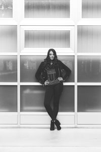 Full length portrait of young woman with hands on hip standing against glass wall