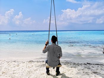 Rear view of man looking at seascape while sitting on swing against sky