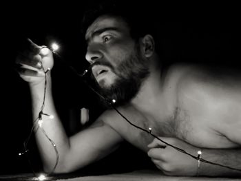 Close-up of shirtless shocked man holding illuminated string lights in darkroom