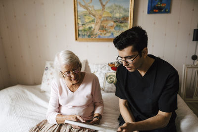 Young male nurse showing mirror to senior woman sitting on bed at home