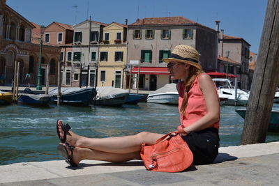 Side view of young woman sitting by canal on sunny day