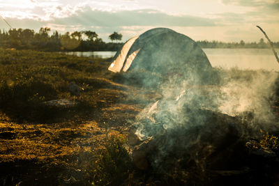 Smoke from campfire by tent at lakeshore during sunset