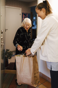 Female caregiver giving grocery bag to senior woman while arriving at home