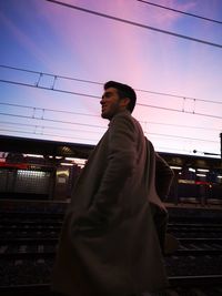 Man standing by railroad tracks against sky during sunset