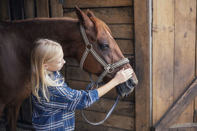 A girl in cowboy clothes in the stable holds a horse by the bridle and stands back to the camera