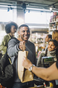 Smiling male customer taking shopping bag from clerk at checkout in convenience store