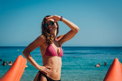 Portrait of woman in bikini standing at beach against blue sky