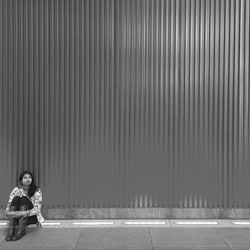 Full length of woman looking away while sitting on footpath by corrugated iron