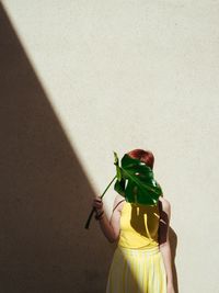 Young woman holding leaf while standing against wall